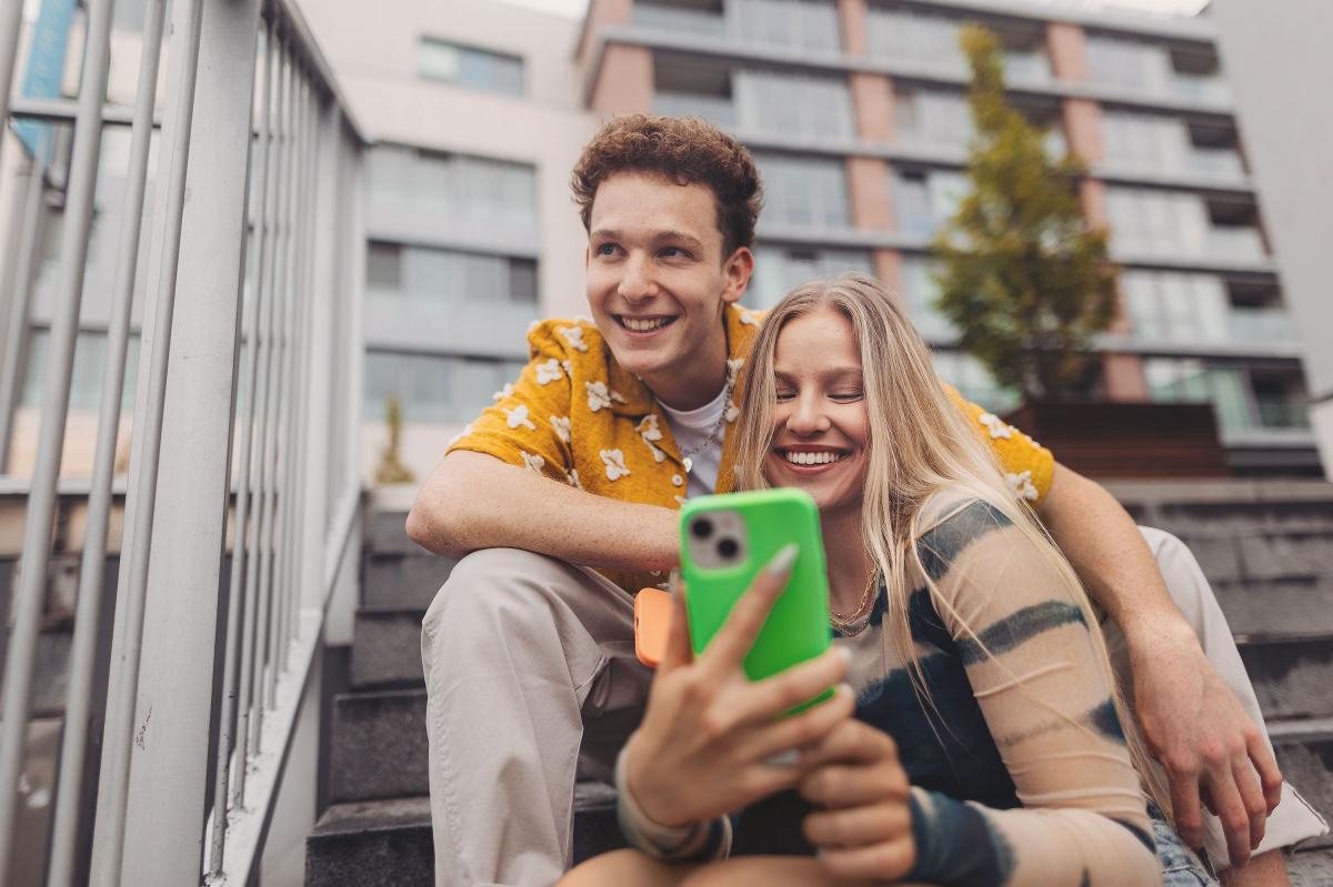 Zwei Teenager sitzen lachend hintereinander. Ein Teenager schaut auf ihr Handy