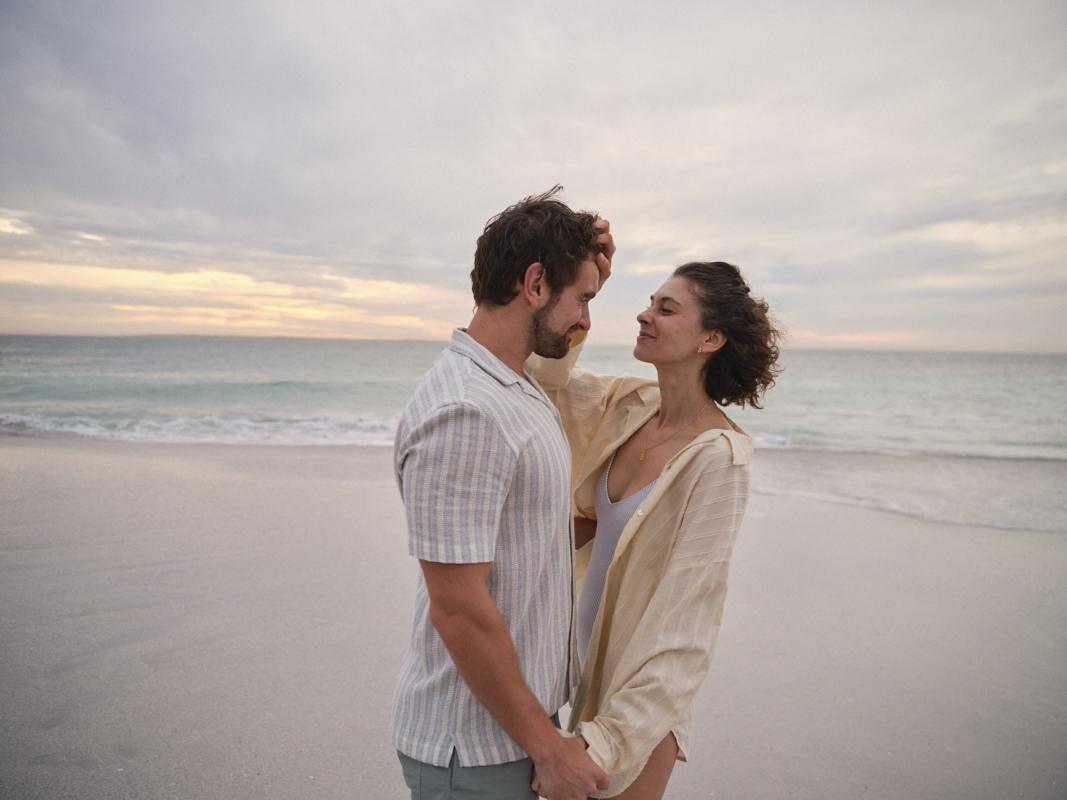 A couple stands on the beach at sunset, gazing at each other with smiles, surrounded by gentle waves and soft, pastel skies.