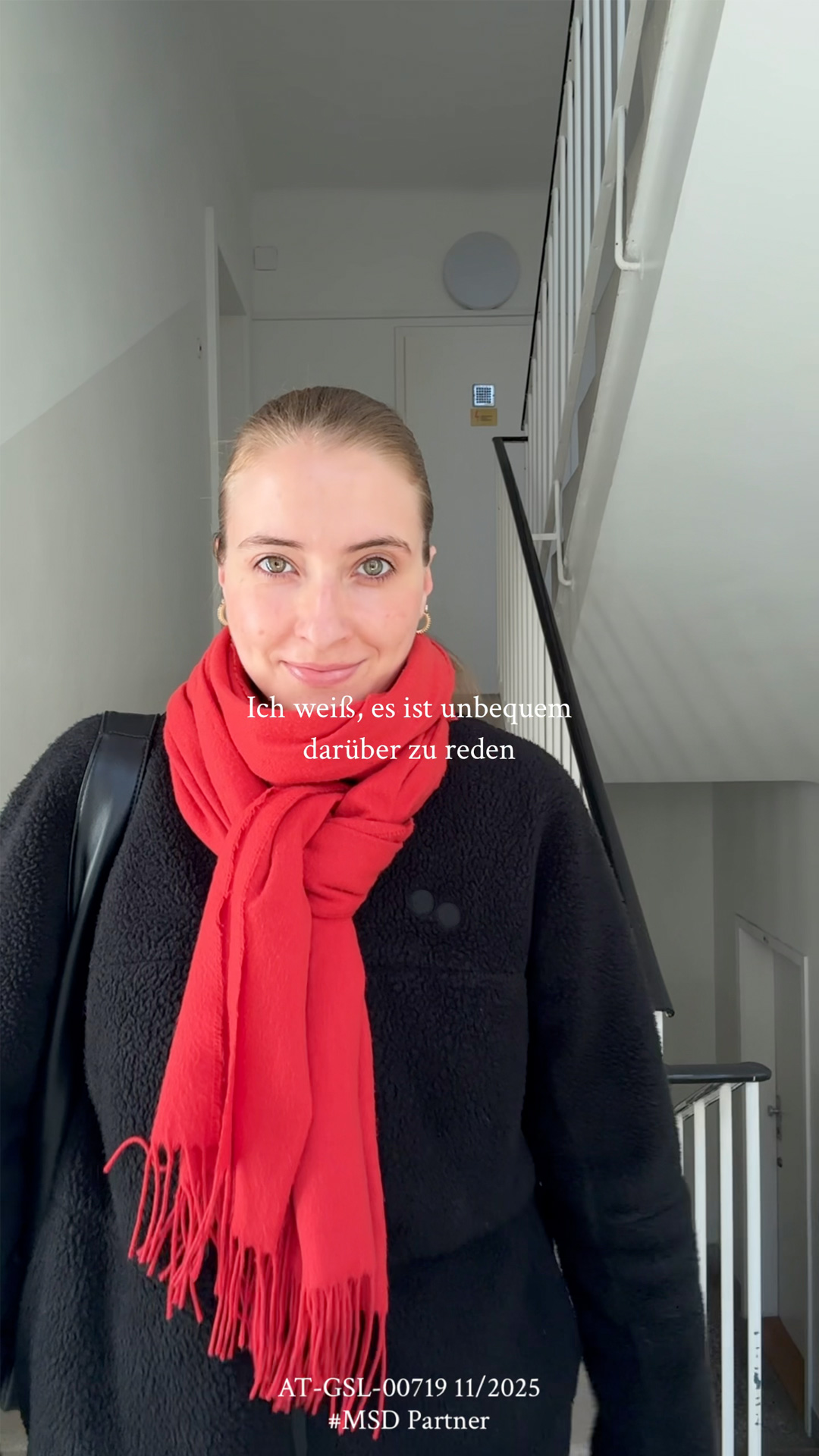 Eine junge Frau mit blondem Haar, rotem Schal und schwarzer Jacke, die an einer Treppe steht, lächelt freundlich.